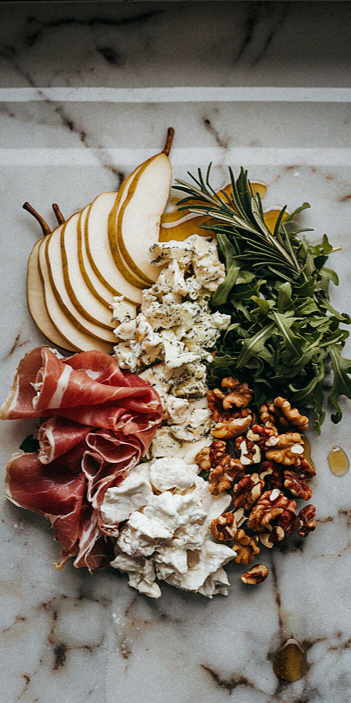 An elegant charcuterie board featuring sliced pears, prosciutto, cheeses, walnuts, arugula, and rosemary on a marble surface.