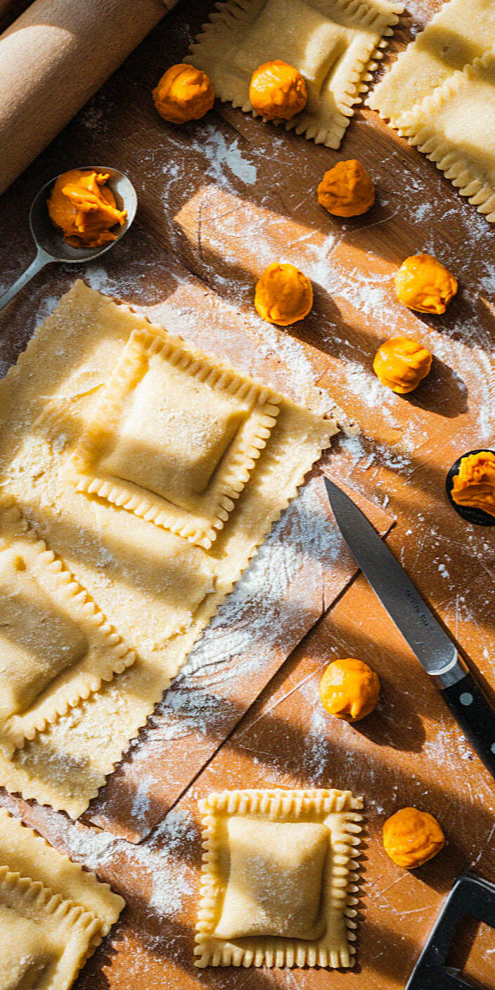 Homemade ravioli being prepared on a floured wooden surface, surrounded by dough, filling, a rolling pin, and a knife.