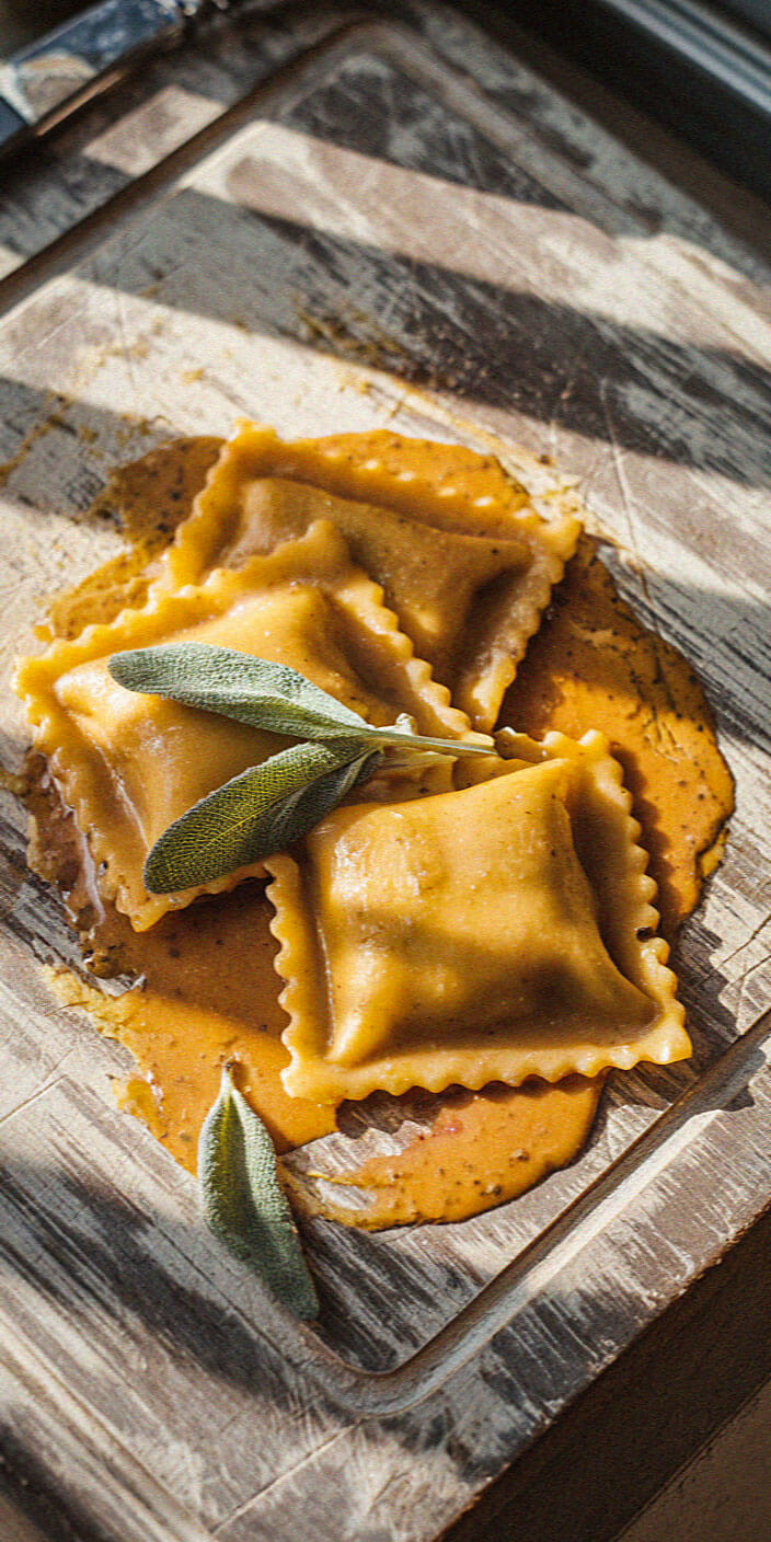 Three ravioli pieces with sage leaves on a wooden board, covered in a creamy orange sauce, lit by natural sunlight.