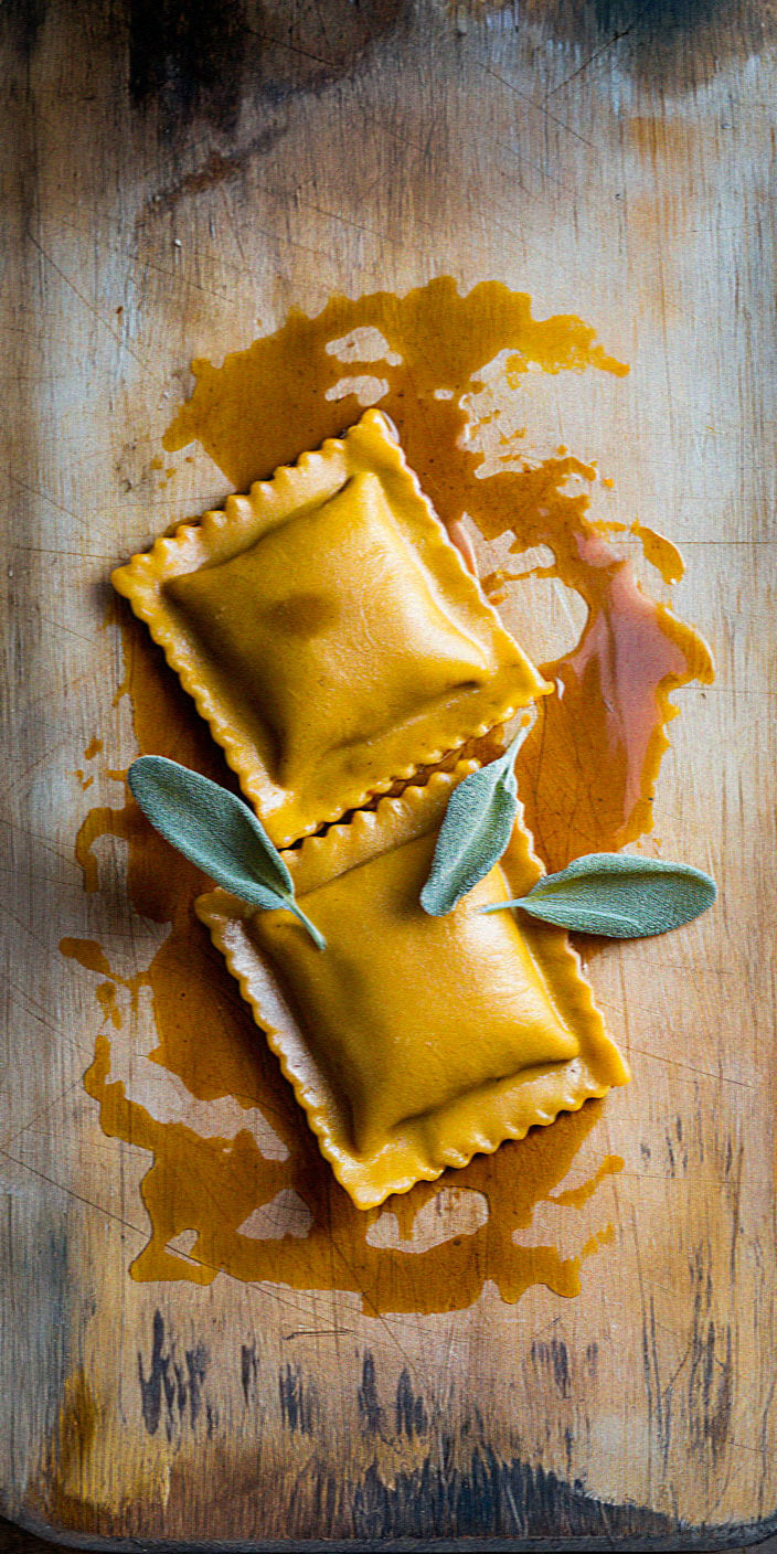 Two pieces of ravioli garnished with sage leaves rest on a wooden cutting board, surrounded by a drizzle of oil.