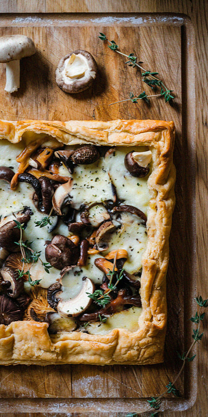 Rustic mushroom tart on wooden board, garnished with thyme. Flaky crust surrounds creamy cheese and assorted mushrooms, creating an appetizing, savory dish presentation.