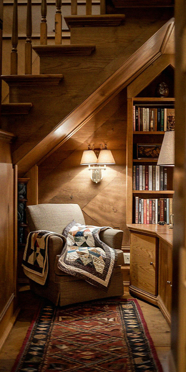 Cozy reading nook under wooden stairs, featuring an armchair with a quilt, bookshelf filled with books, and warm lighting from wall lamp.