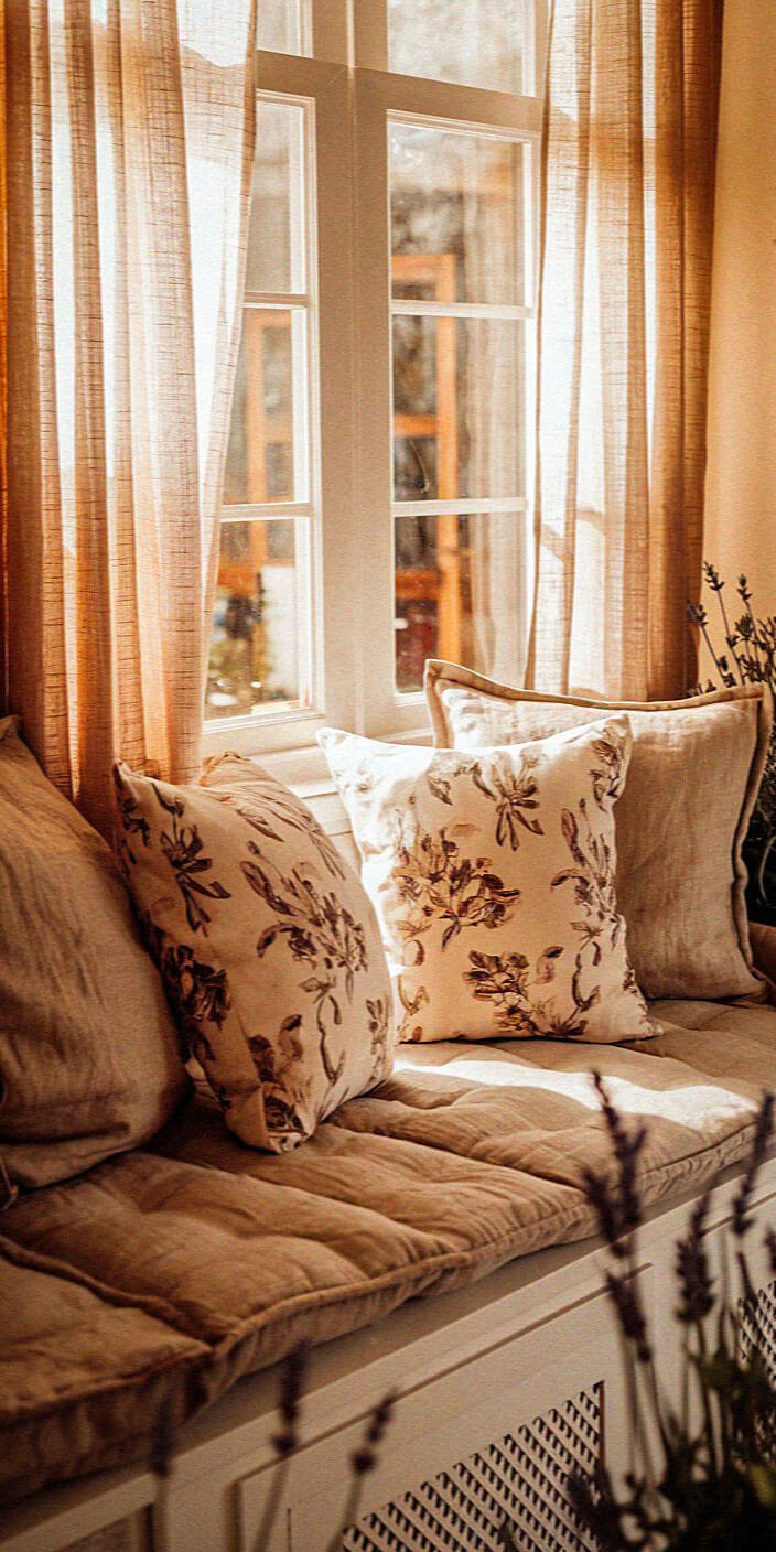 Cozy window seat with floral cushions, soft beige tones, and sheer curtains letting in warm sunlight. Peaceful and inviting atmosphere.