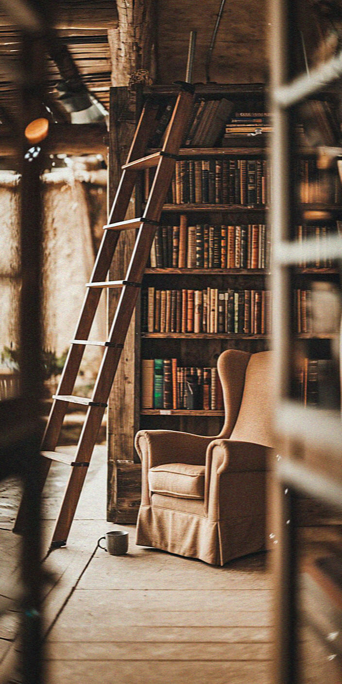 Cozy library corner with a tall bookshelf, wooden ladder, plush armchair, and a mug on the floor, evoking a warm, inviting atmosphere.