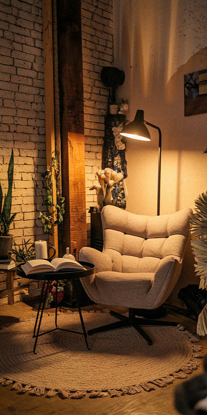 Cozy corner with an armchair, open book on a table, and soft lighting, surrounded by plants and rustic brick walls.