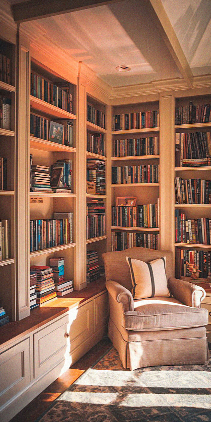 Cozy library corner with a beige armchair, surrounded by bookshelves filled with colorful books. Sunlight streams through a window, creating warmth.