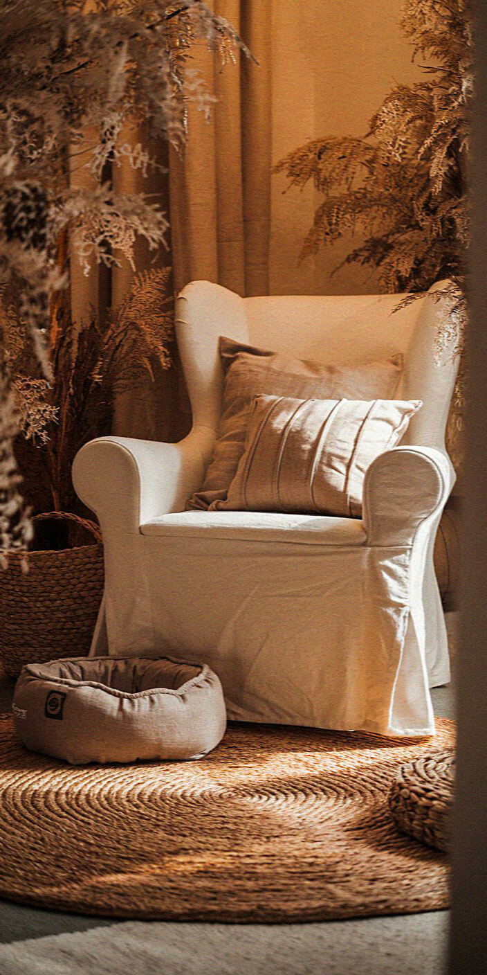 Cozy corner with a white armchair, beige cushions, a round woven rug, dog bed, and decorative plants in a warm, serene ambiance.