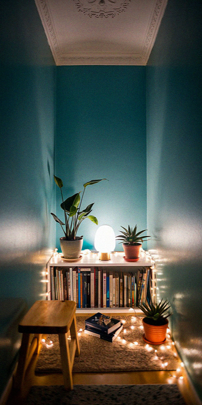 Cozy reading nook with a small wooden stool, bookshelf, potted plants, soft lighting, and blue walls, creating a calming atmosphere.