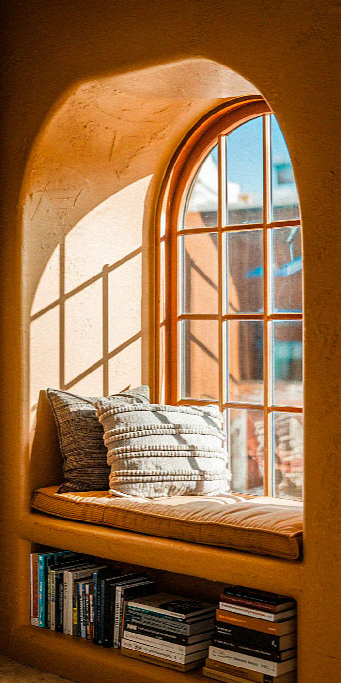Cozy nook with arched window, sunlight streaming in, cushioned seat, and bookshelves filled with various books, creating a warm, inviting atmosphere.