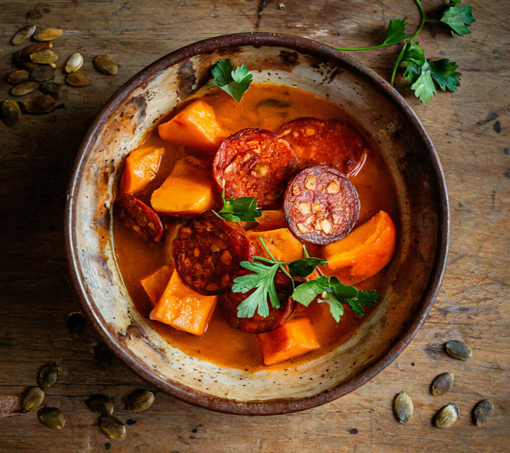 A rustic bowl with sliced chorizo, sweet potato, and parsley garnish on a wooden surface with scattered seeds.
