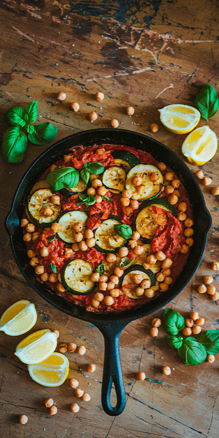 A skillet with tomatoes, zucchini, and chickpeas on a rustic wooden table, garnished with fresh basil and lemon wedges.