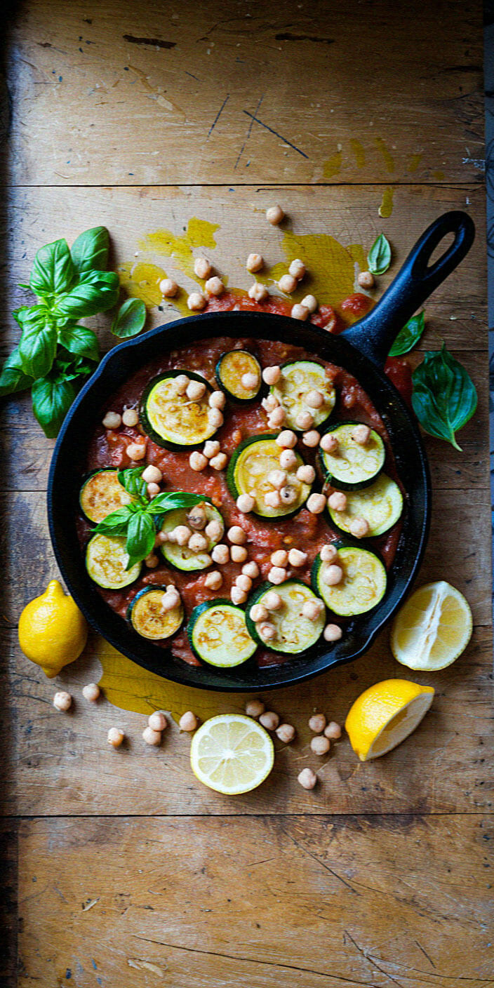 Skillet with zucchini, chickpeas, and tomato sauce on wooden surface, garnished with basil and lemon halves. Vibrant and colorful presentation.