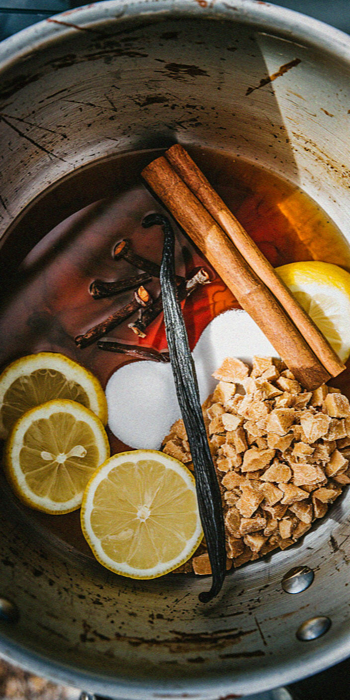 A pot with lemon slices, cinnamon sticks, cloves, rock sugar, vanilla bean, and liquid, creating a spiced aromatic mixture.