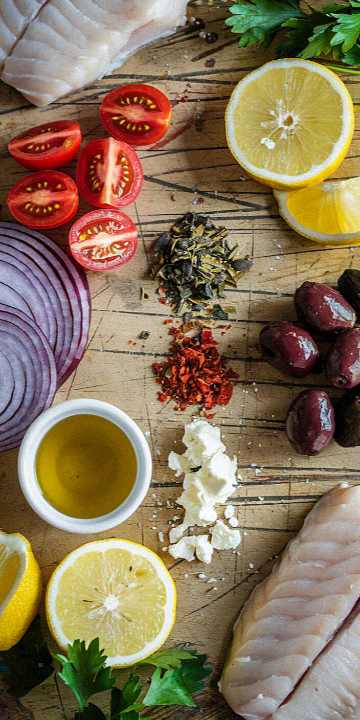 Ingredients on a wooden board: fish fillets, lemon slices, tomatoes, olives, red onion, olive oil, herbs, spices, cheese, and parsley.
