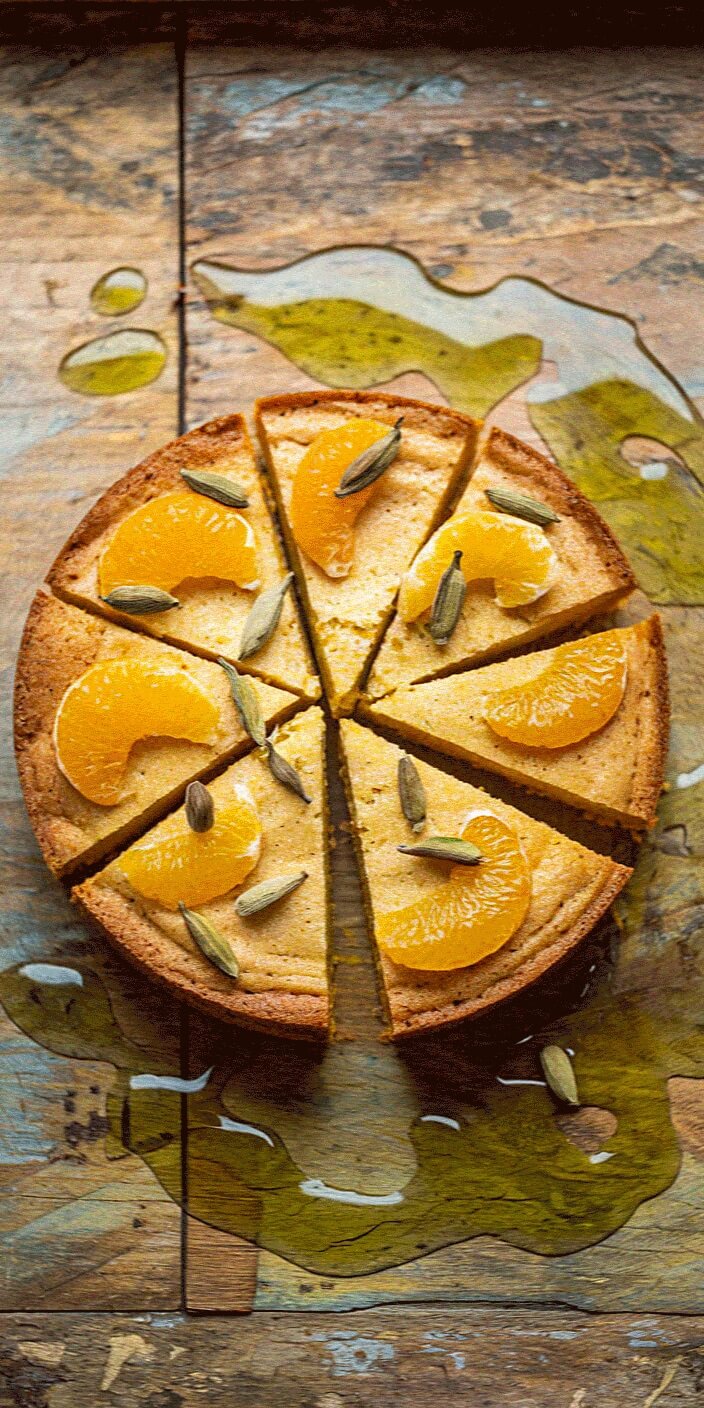 A sliced cake topped with orange segments and cardamom sits on a wooden surface with olive oil drizzled around it.