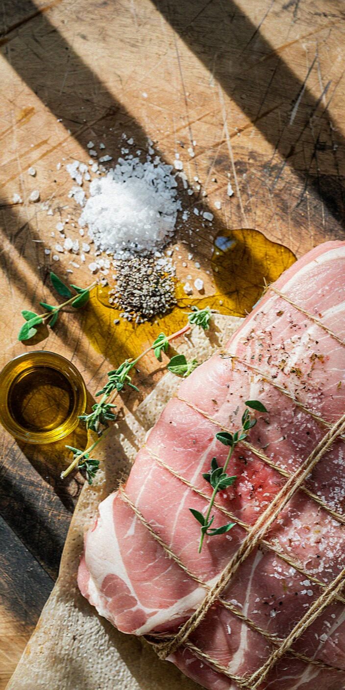 Raw pork roast tied with twine, accompanied by coarse salt, black pepper, olive oil, and fresh herbs on a wooden surface, illuminated by sunlight.