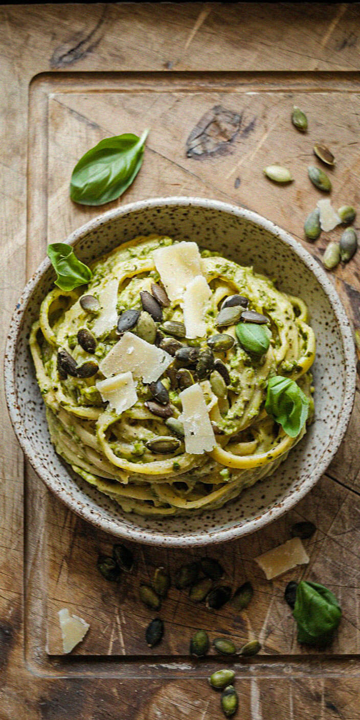 A bowl of pasta with pesto sauce, topped with parmesan, pumpkin seeds, and fresh basil leaves on a wooden surface.