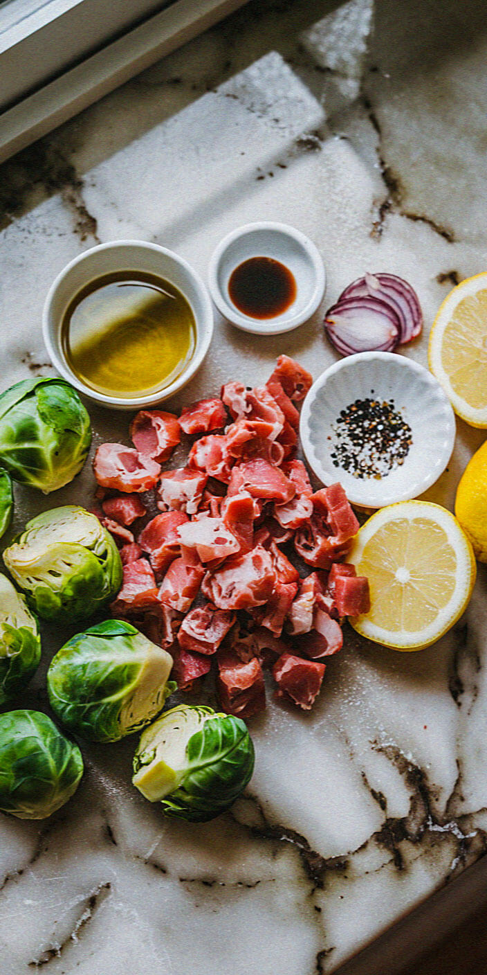 Fresh Brussels sprouts, diced meat, lemon halves, oil, vinegar, onion slices, and spices are arranged on a marble surface near a window.