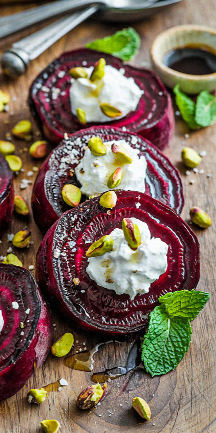 Sliced roasted beets topped with creamy cheese, pistachios, and fresh mint leaves on a wooden board, accompanied by a small bowl of balsamic vinegar.