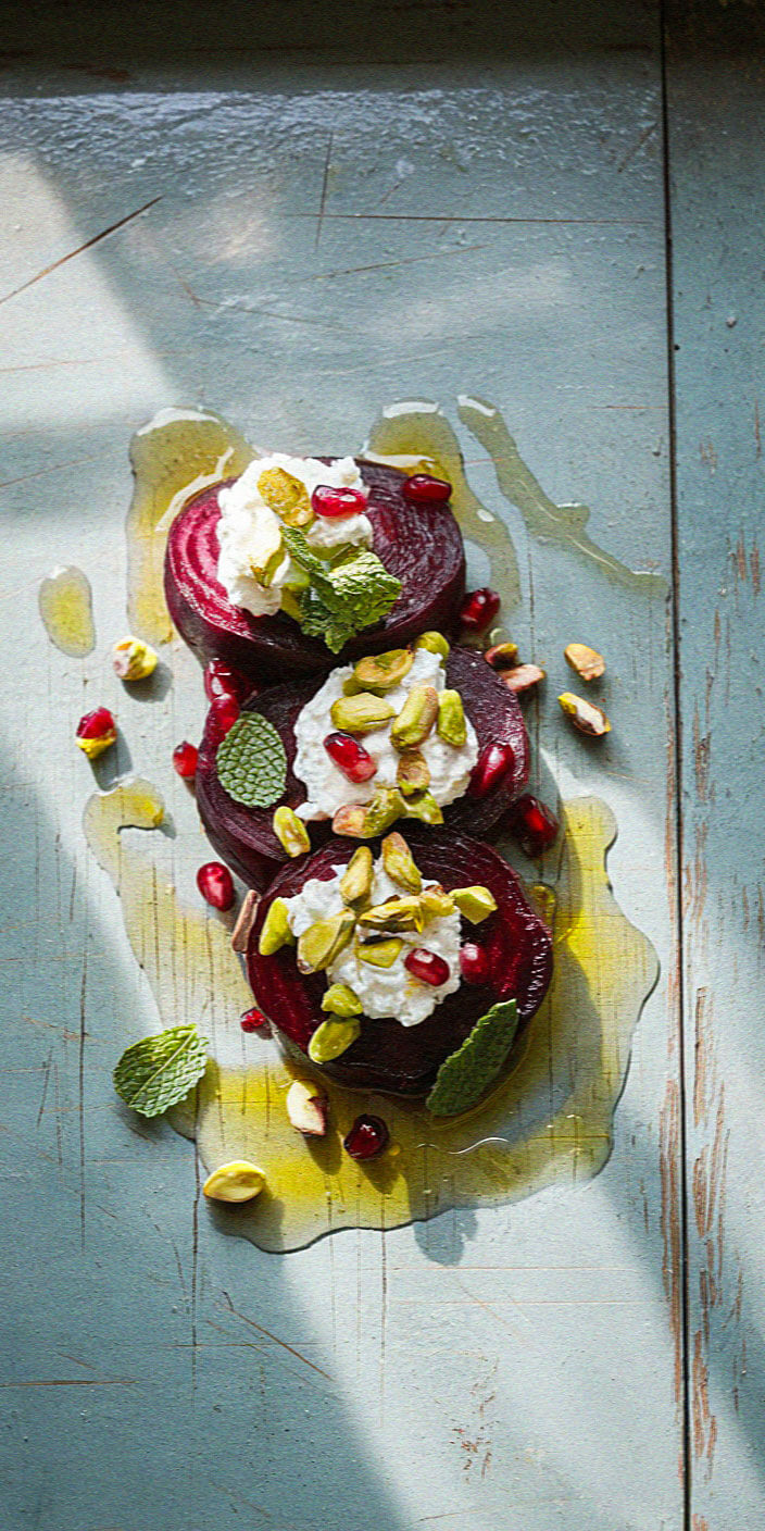 Sliced beets topped with cheese, pistachios, pomegranate seeds, and mint leaves on a rustic, blue wooden surface, drizzled with olive oil.