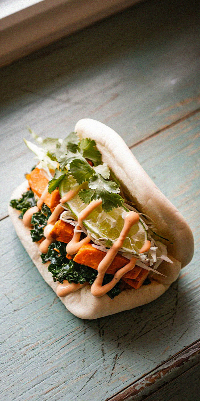A bao bun filled with sweet potato, kale, lime, cilantro, and drizzled sauce on a rustic wooden surface, creating a colorful, appetizing display.