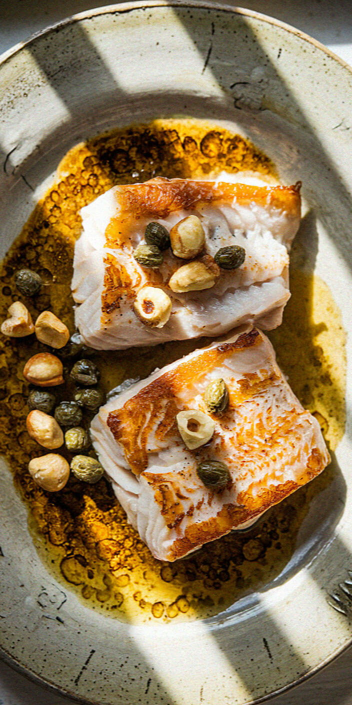 Two pieces of cooked fish with capers and nuts on a plate, drizzled with golden oil, in natural light.