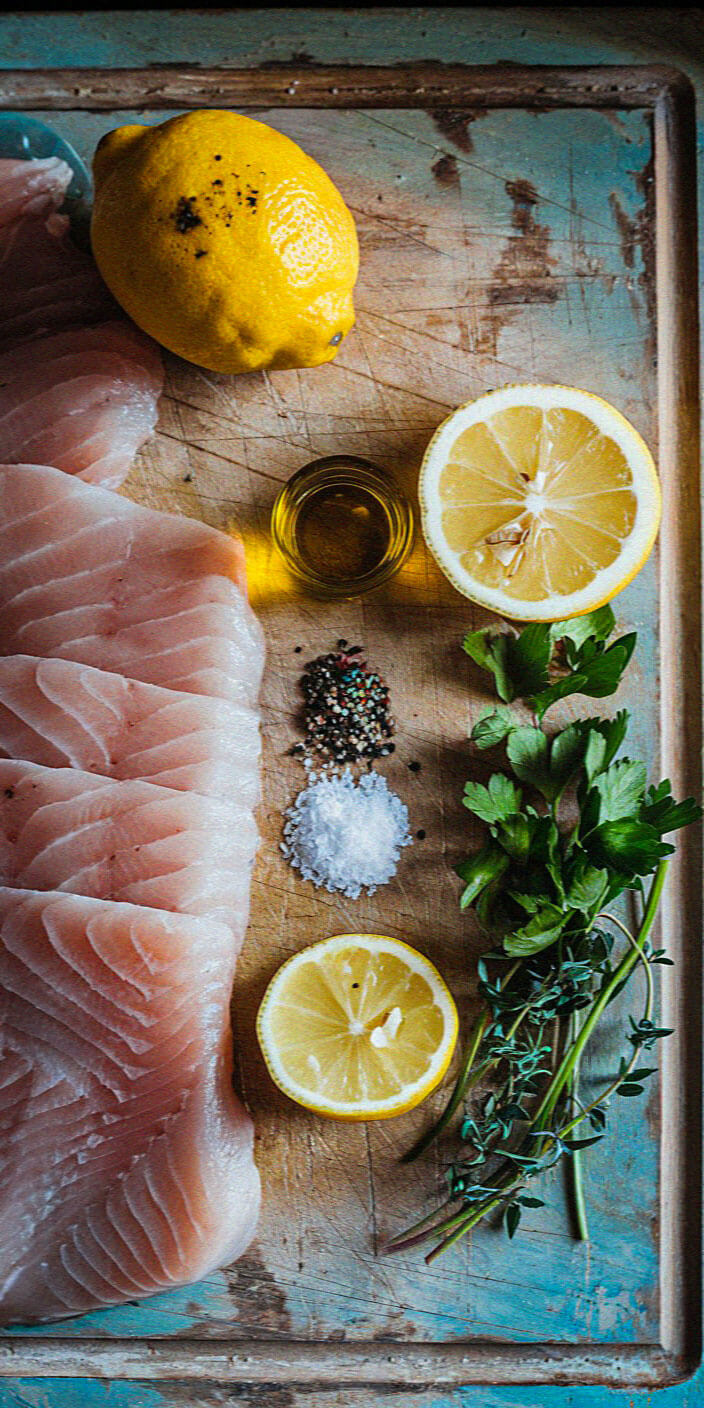 Raw fish fillets, lemon halves, herbs, salt, pepper, and olive oil arranged on a rustic wooden board, ready for preparation.