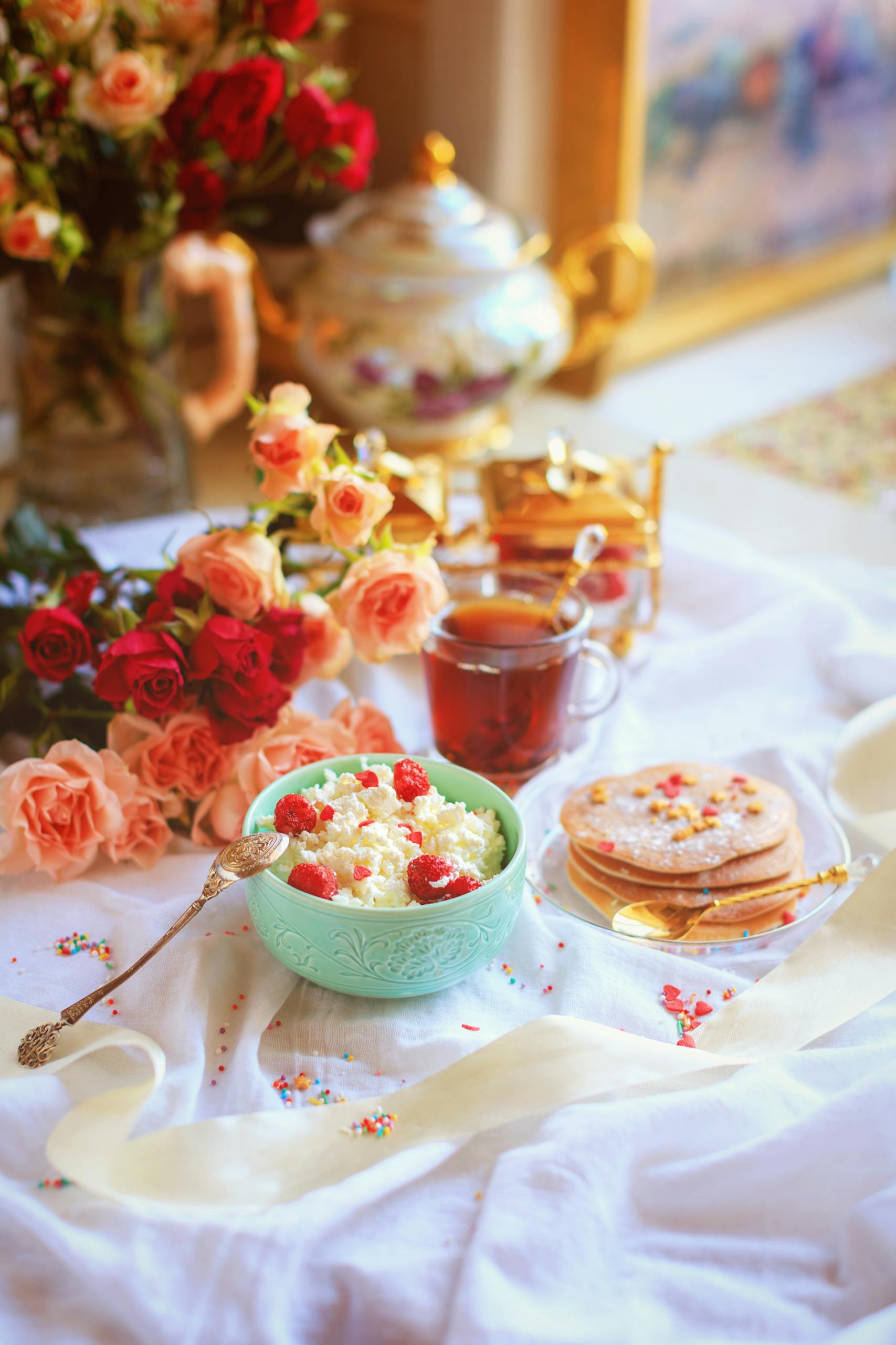 A romantic breakfast scene featuring a bowl of porridge, pancakes, and roses beside a cup of tea.