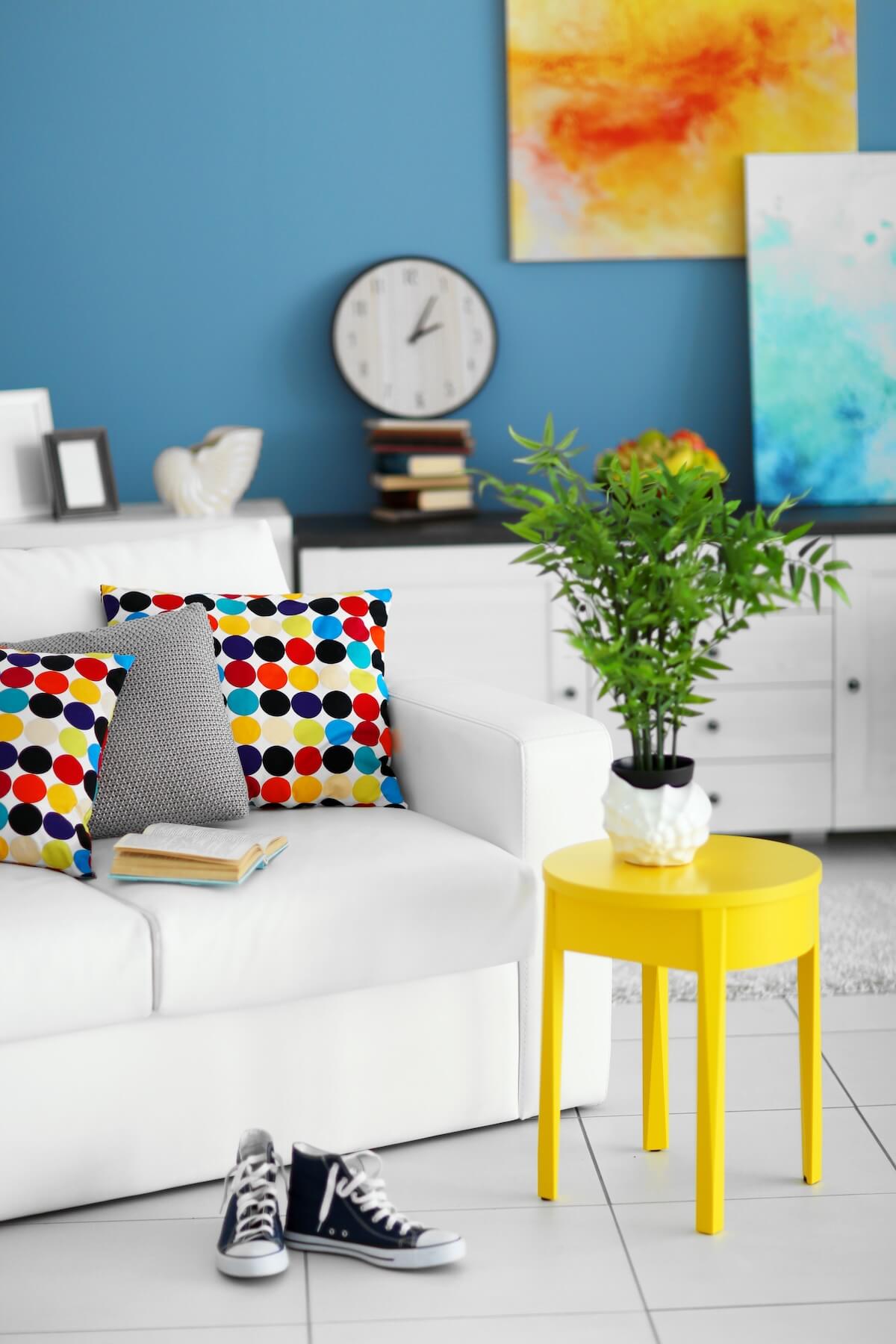 Modern living room with a blue wall, dotted pillows on a white sofa, art, clock, yellow table, sneakers, books, and a plant.
