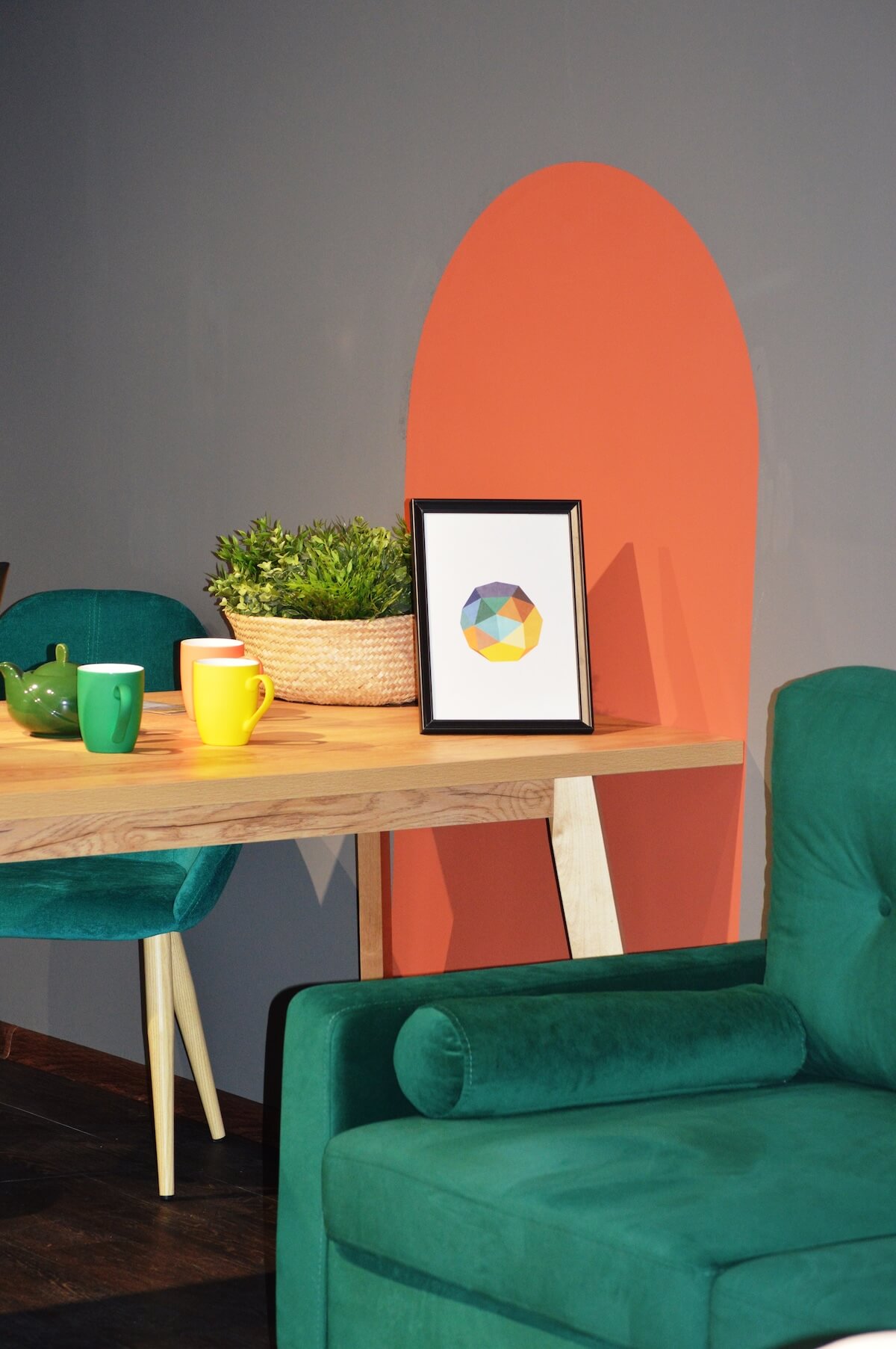A modern room with green chairs, a wooden table, colorful mugs, framed geometric artwork, basket of greenery, and an orange wall accent.