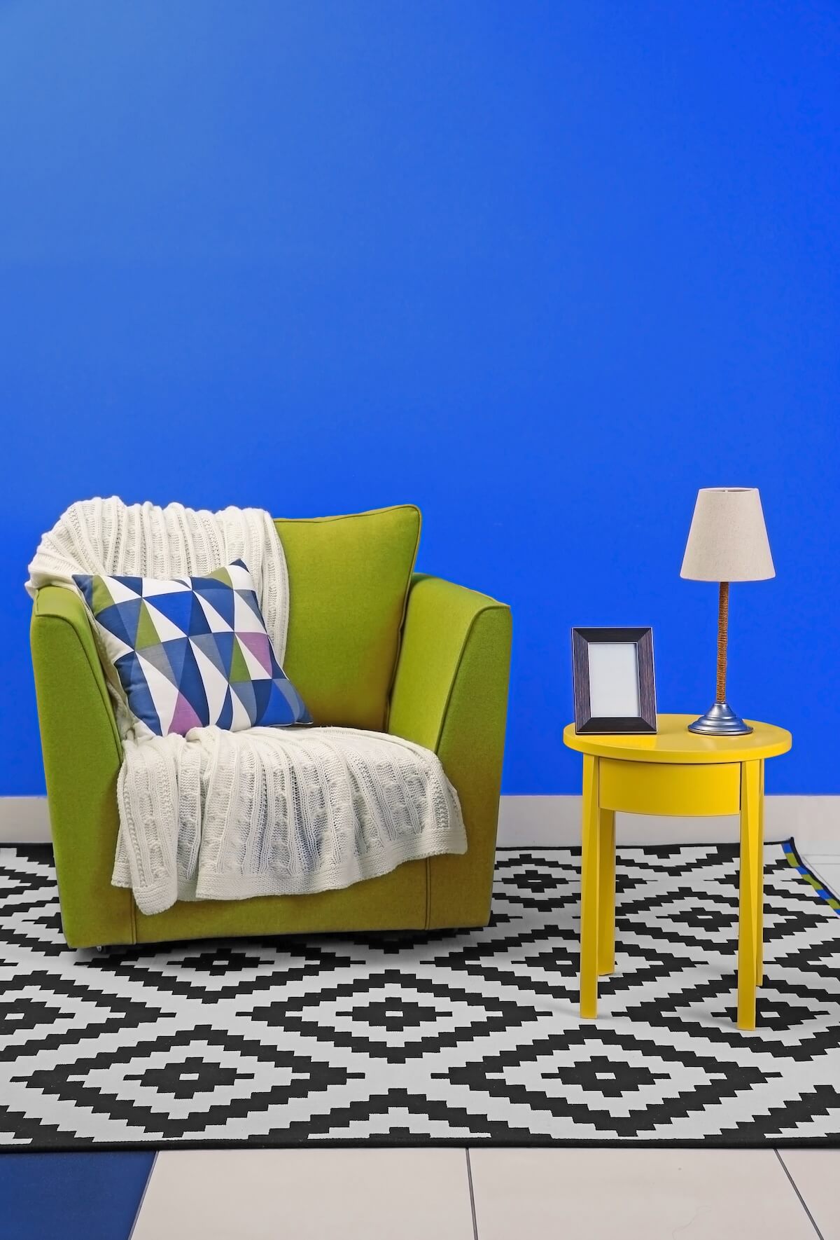 A bright room features a green armchair with a geometric-patterned pillow and a knitted throw, beside a yellow table with a lamp.