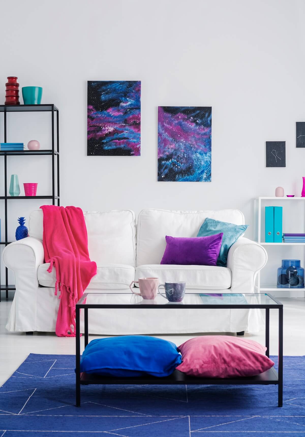 Modern living room features a white sofa with colorful cushions and blanket, abstract art, glass coffee table, and minimalist shelving with vibrant decor.