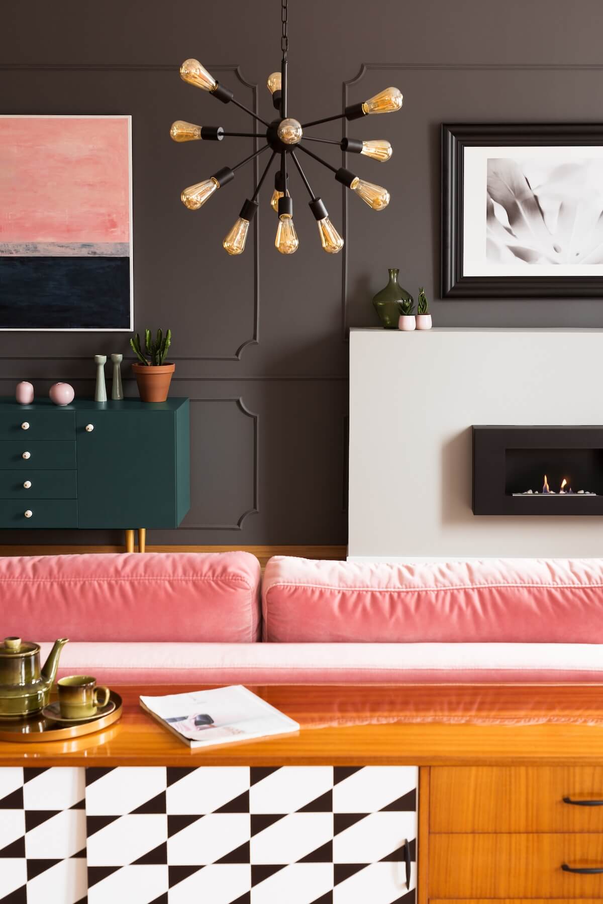 Modern living room with geometric patterns, pink couch, abstract art, and sputnik chandelier. Decor includes green cabinet, fireplace, and potted plants.