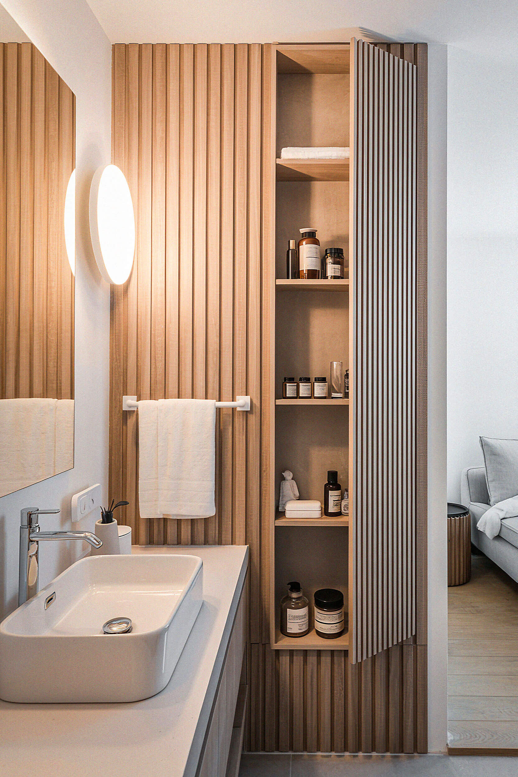 Modern bathroom featuring a vertical wood slat wall, a white vanity sink, a round wall light, and tall open shelves with toiletries.