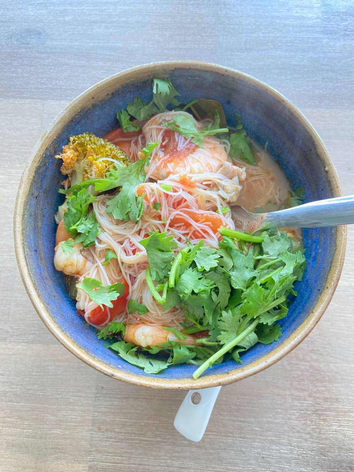 A blue ceramic bowl of noodle soup topped with shredded chicken, fresh cilantro, and greens, with a spoon resting inside.